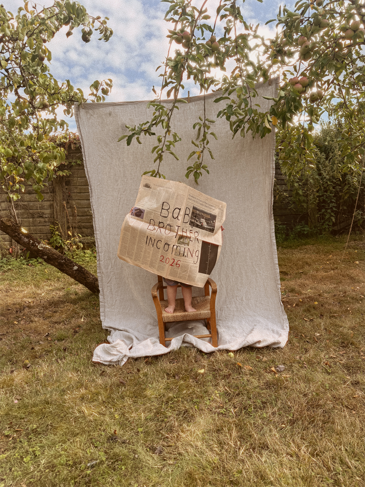 Person sitting on a chair holding a newspaper with text, standing behind a white curtain in an outdoor setting.