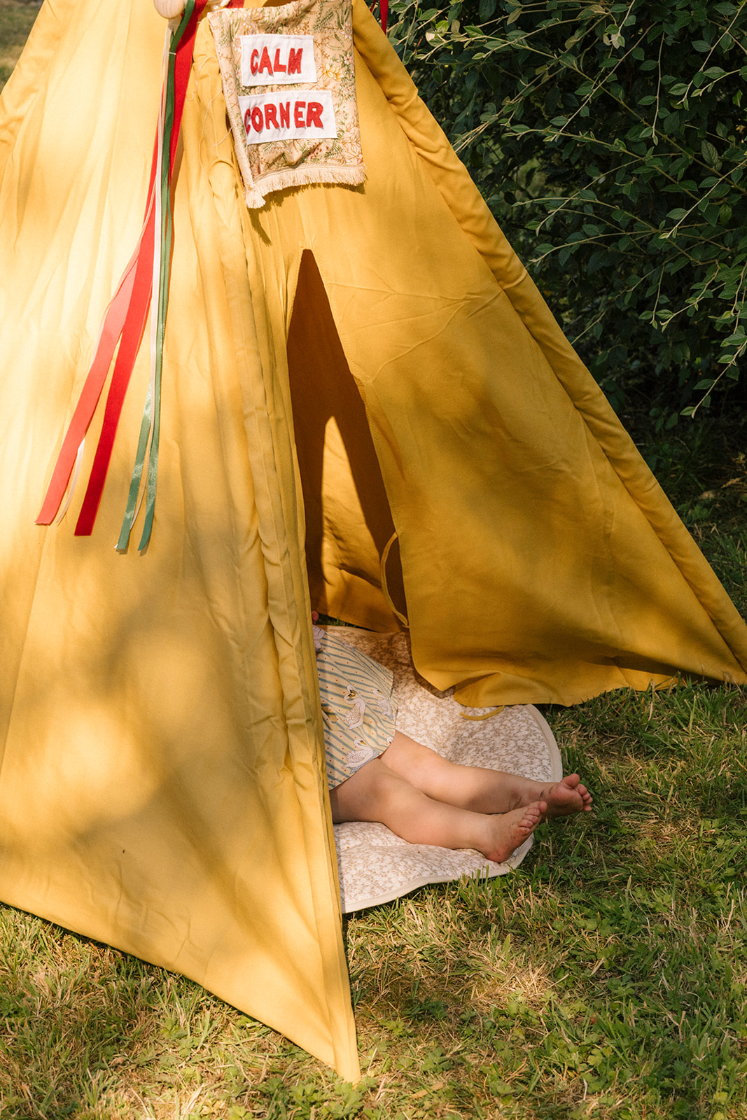 Person peeking out from a yellow tent with 'CALM CORNER' sign, surrounded by greenery