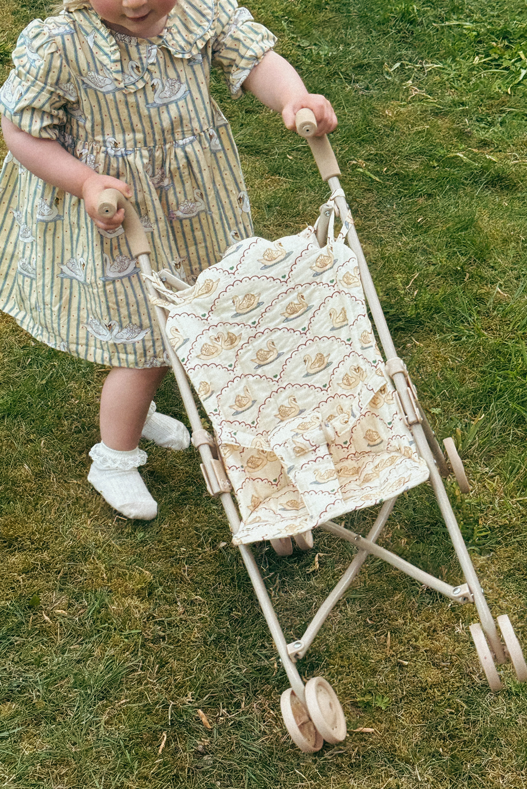 Child in a vintage dress pushing a matching vintage-style doll stroller on grass.