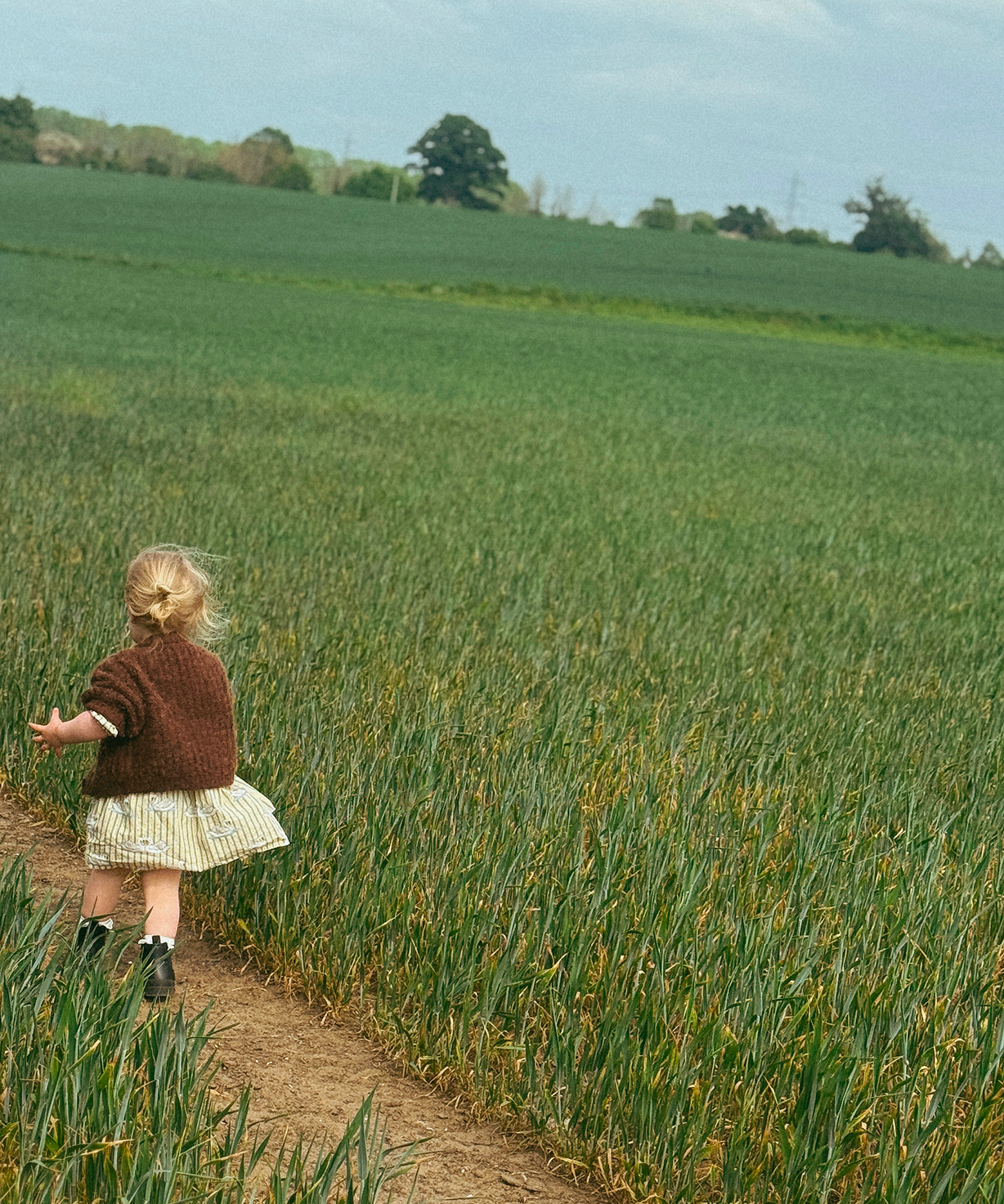 Child walking along a dirt path through a green field with a clear sky.