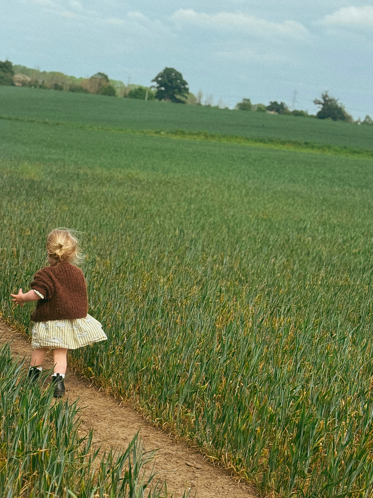 Child walking along a dirt path through a green field with a clear sky.