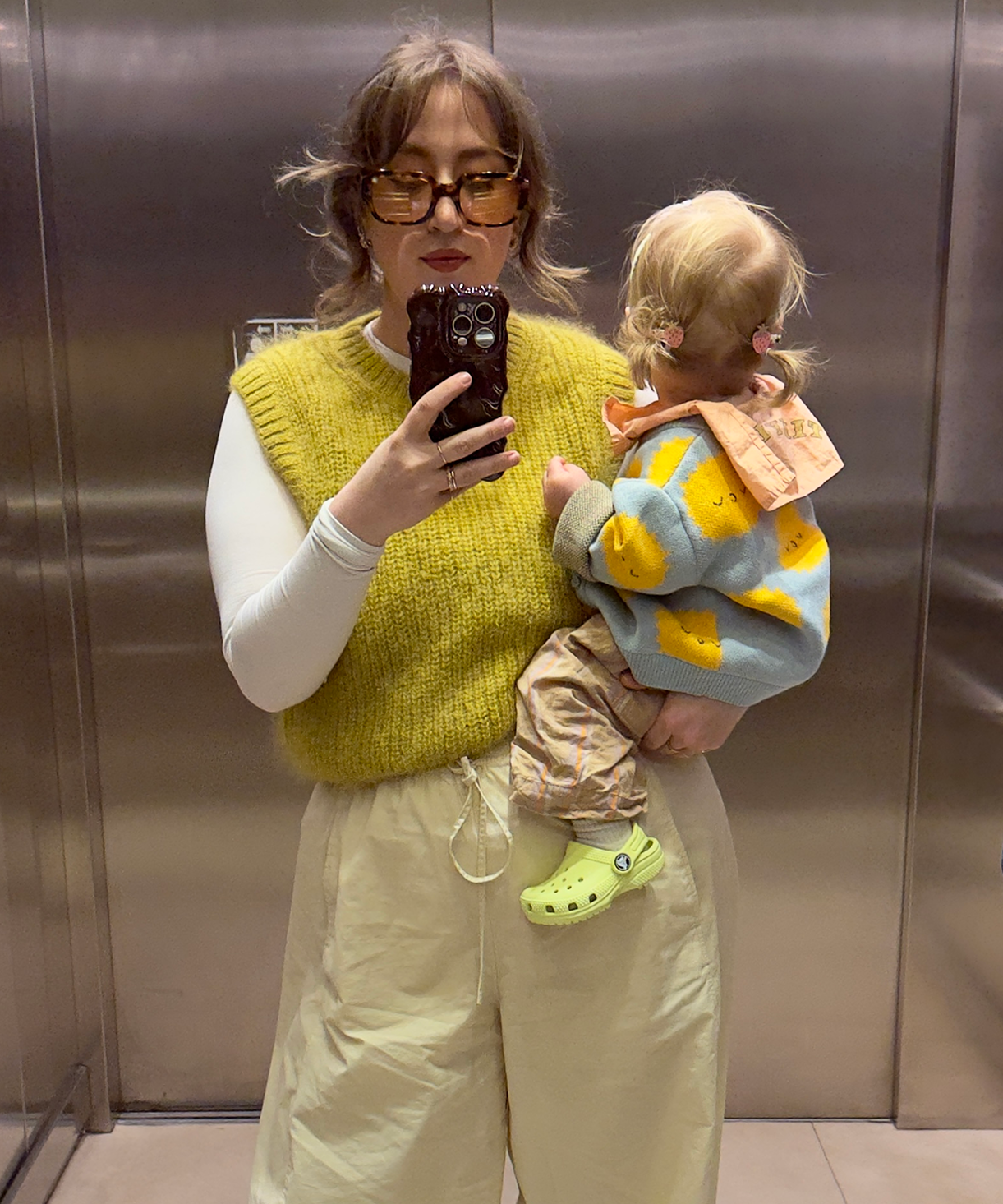 Woman taking a mirror selfie with a child wearing a colorful sweater in an elevator.