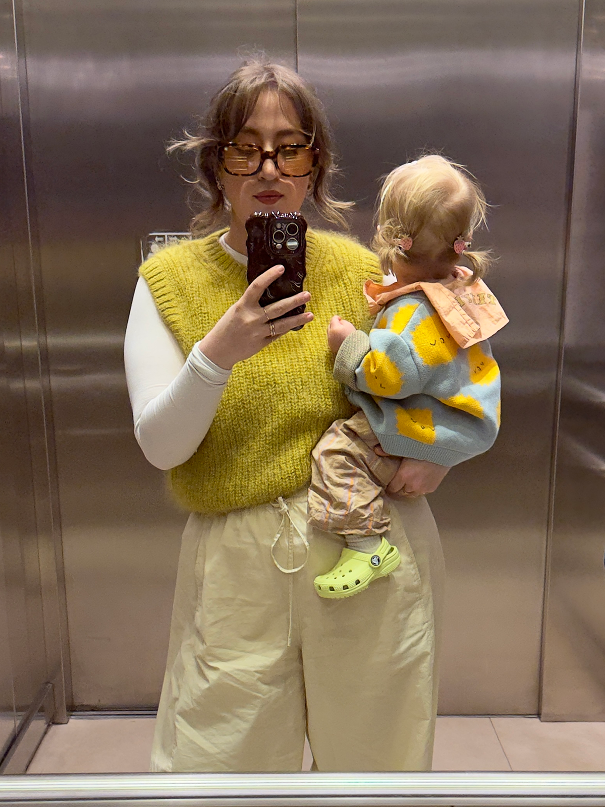 Woman taking a mirror selfie with a child wearing a colorful sweater in an elevator.