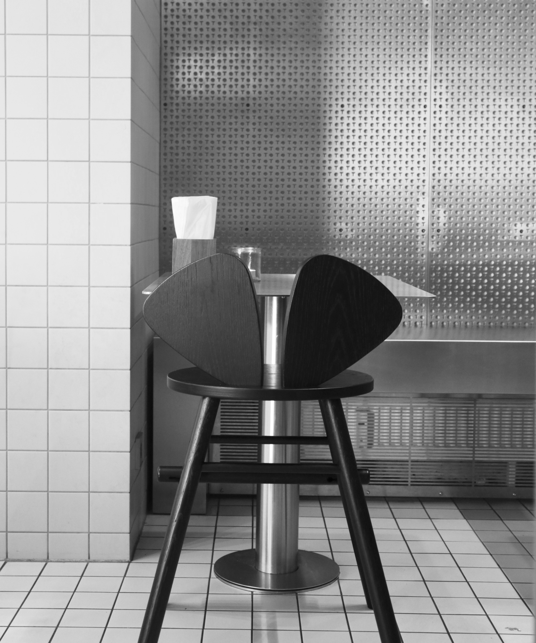 A black modern bar stool with a unique curved backrest, positioned next to a small round table in a minimalist, black-and-white tiled restaurant or cafe setting.