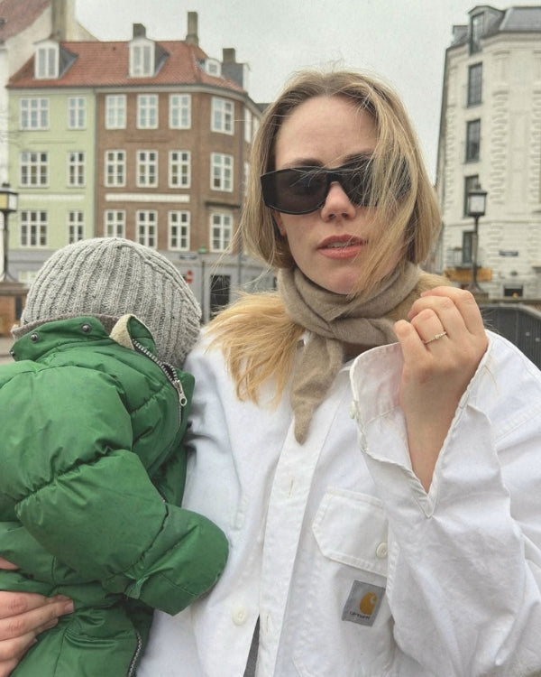Woman in a white coat and sunglasses holding a child in a green coat, with a cityscape in the background.