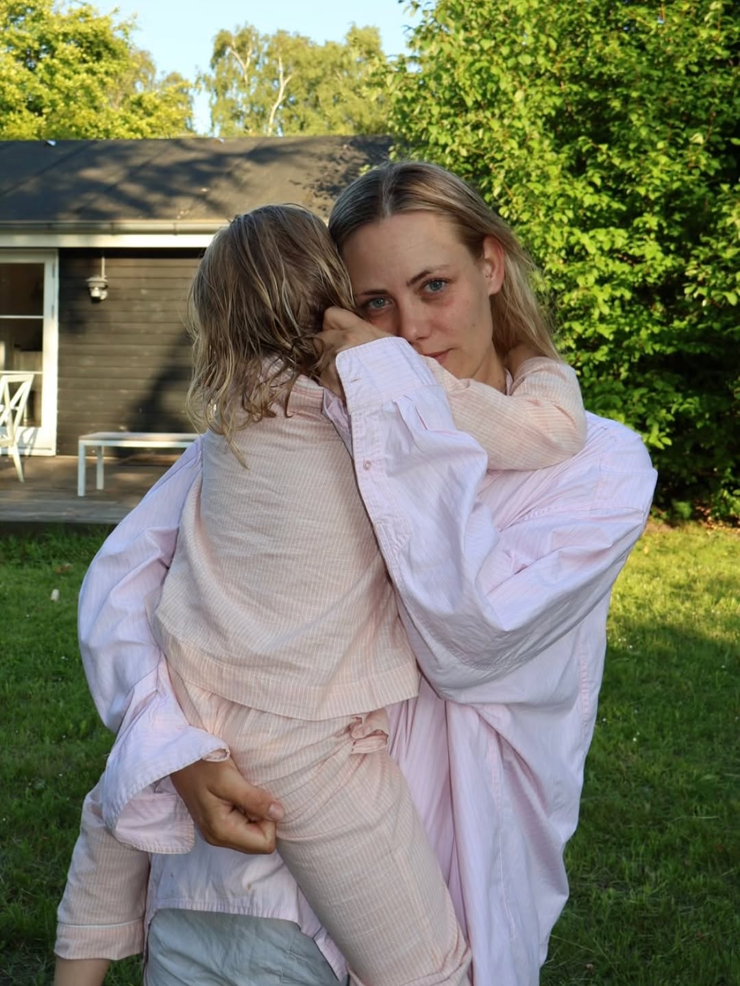 Woman holding a child outdoors with a house and trees in the background