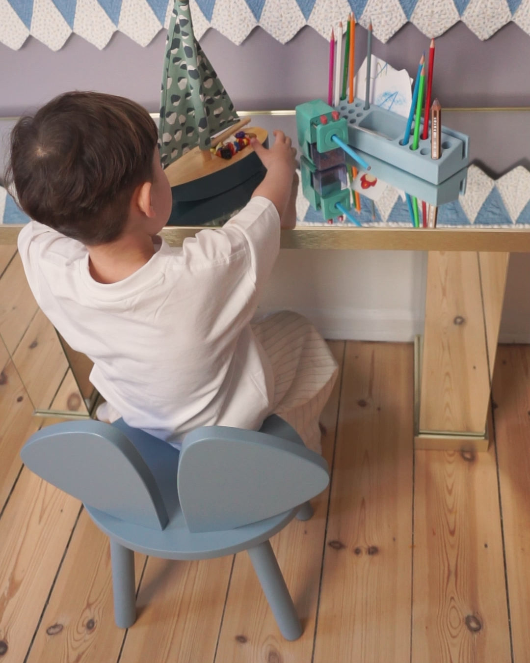 Child seated on a blue Nofred Mouse Chair, playing with a toy boat at a wooden table with coloured pencils.