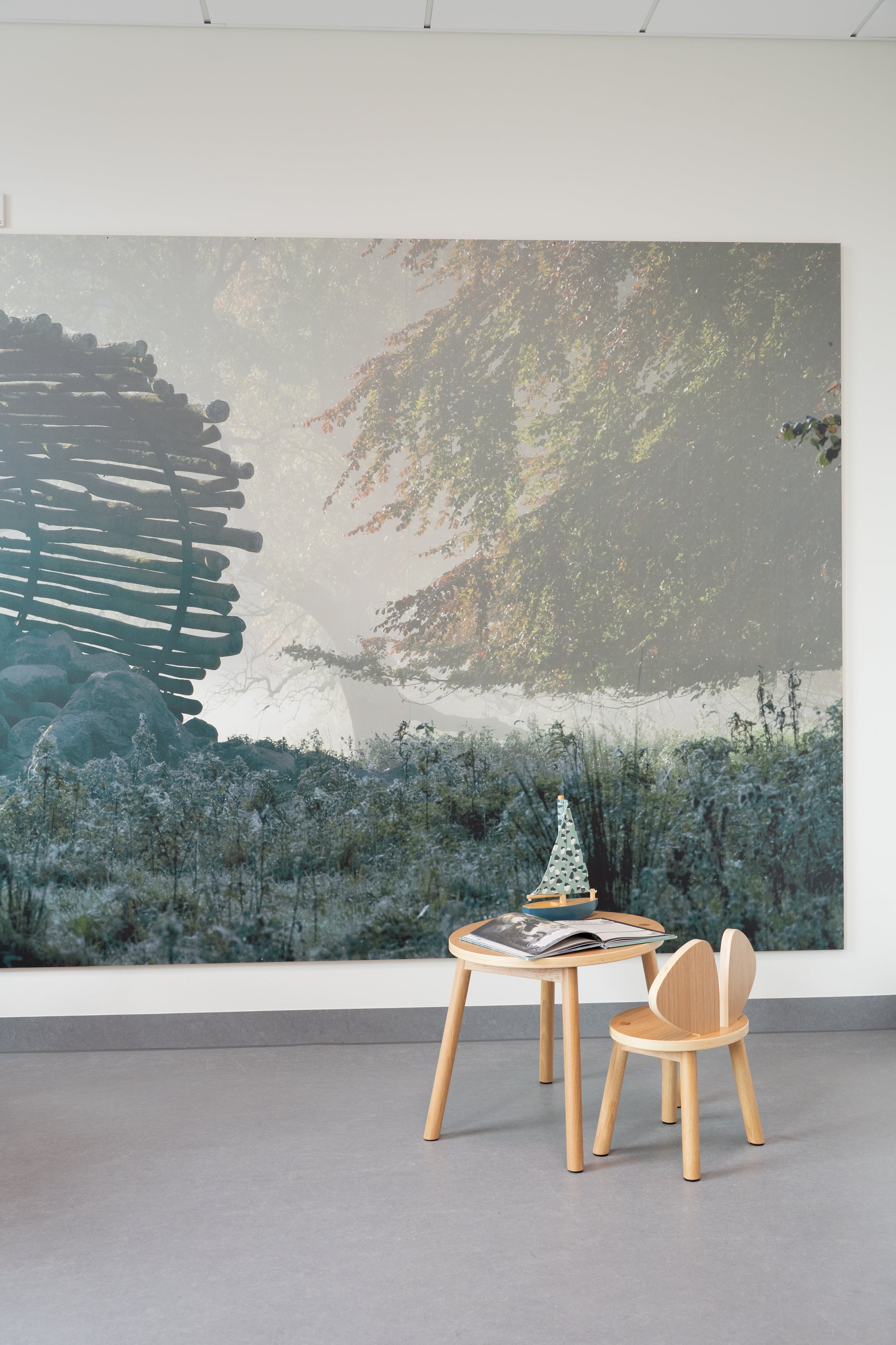 Room with a large wall mural of a natural landscape, featuring trees and a lake, with a small wooden table and chair in front.