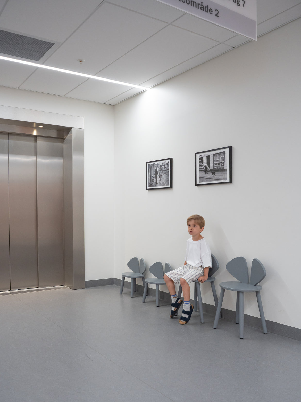 Child sitting on a chair in a waiting room with an elevator and framed pictures on the wall.