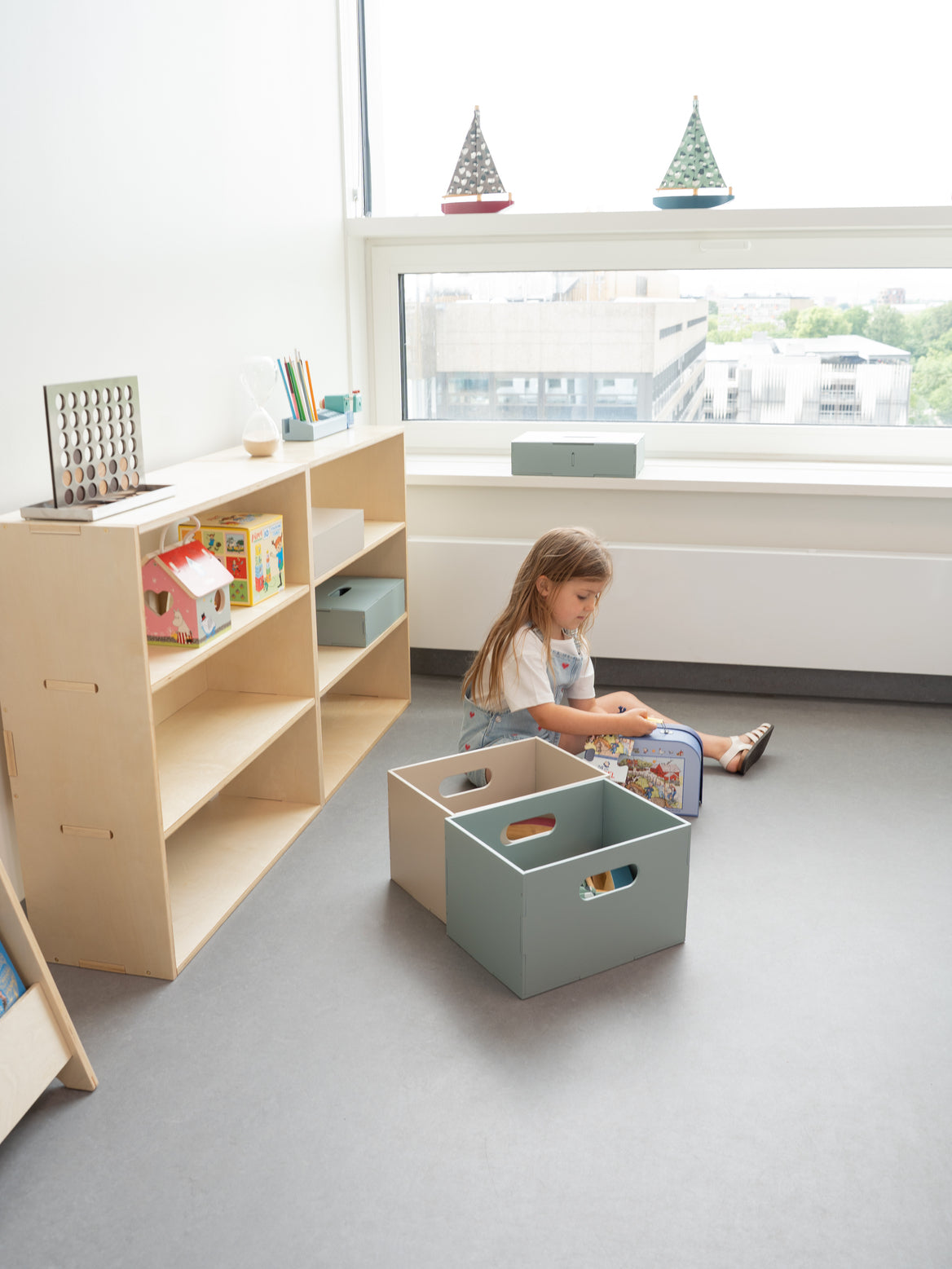 Child playing with toys in a storage box in a room with shelves and large windows.
