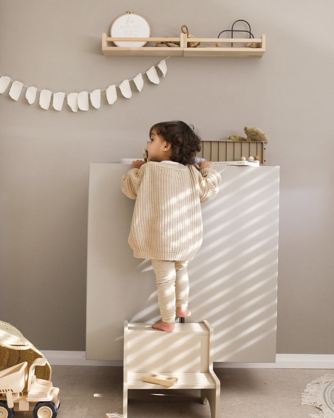 Child standing on a small stool against a white wall with toys and shelves in the background.