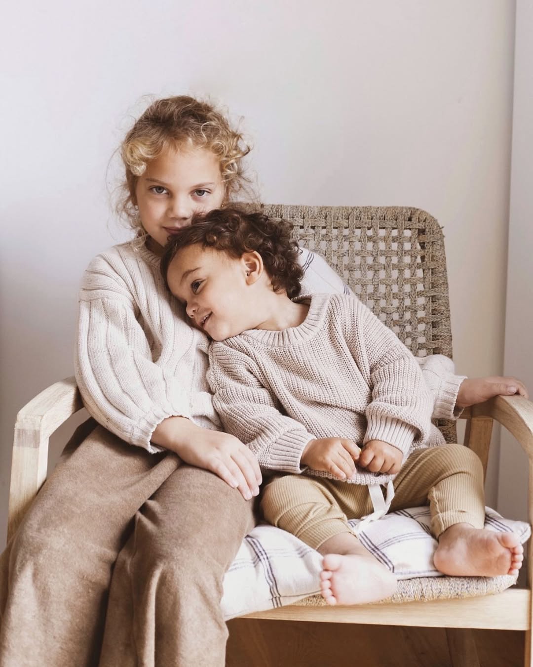 Two children sitting on a wicker chair, one embracing the other, in a cozy indoor setting.