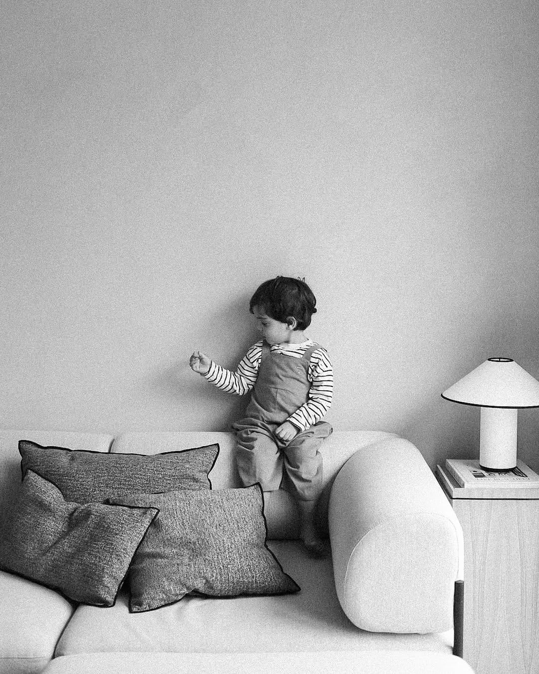 Child standing on a sofa with cushions, next to a lamp on a side table against a plain wall.