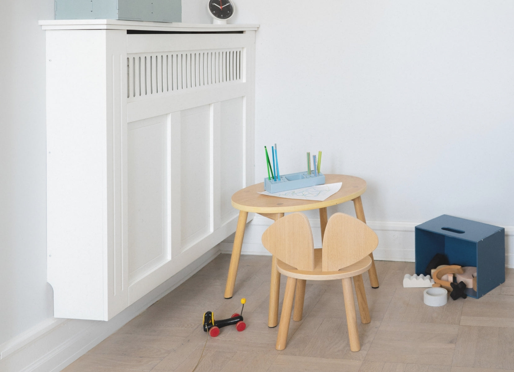 Children's wooden table and chairs with toys on a light wood floor against a white wall.