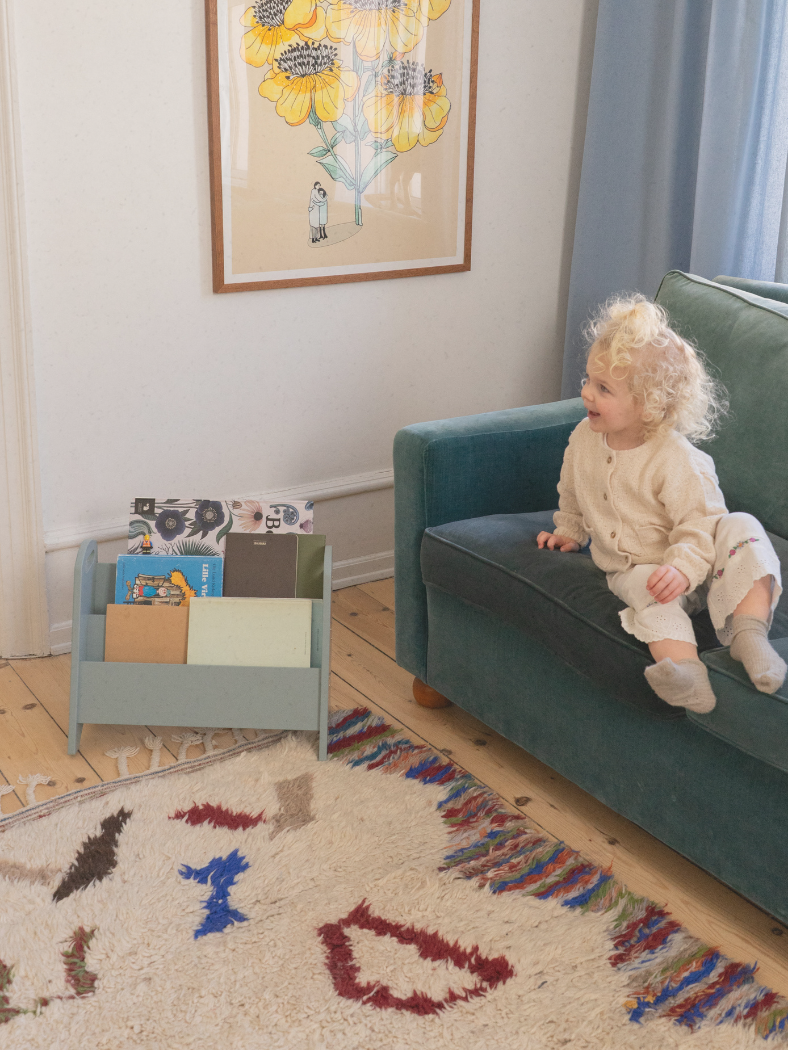 Child sitting on a green couch in a room with a colorful rug and bookshelf.