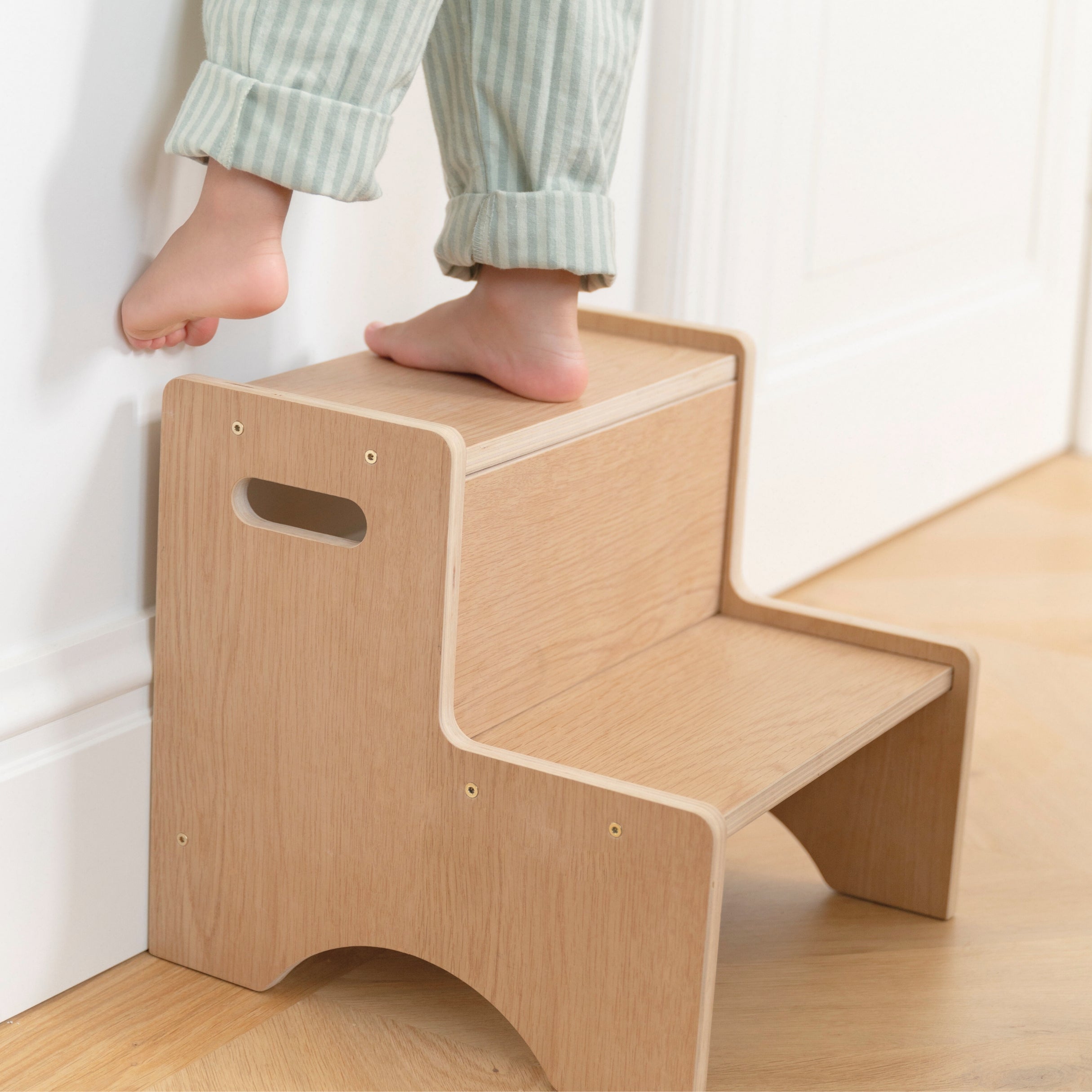 Wooden step stool with a child's feet on it, on a light wooden floor.