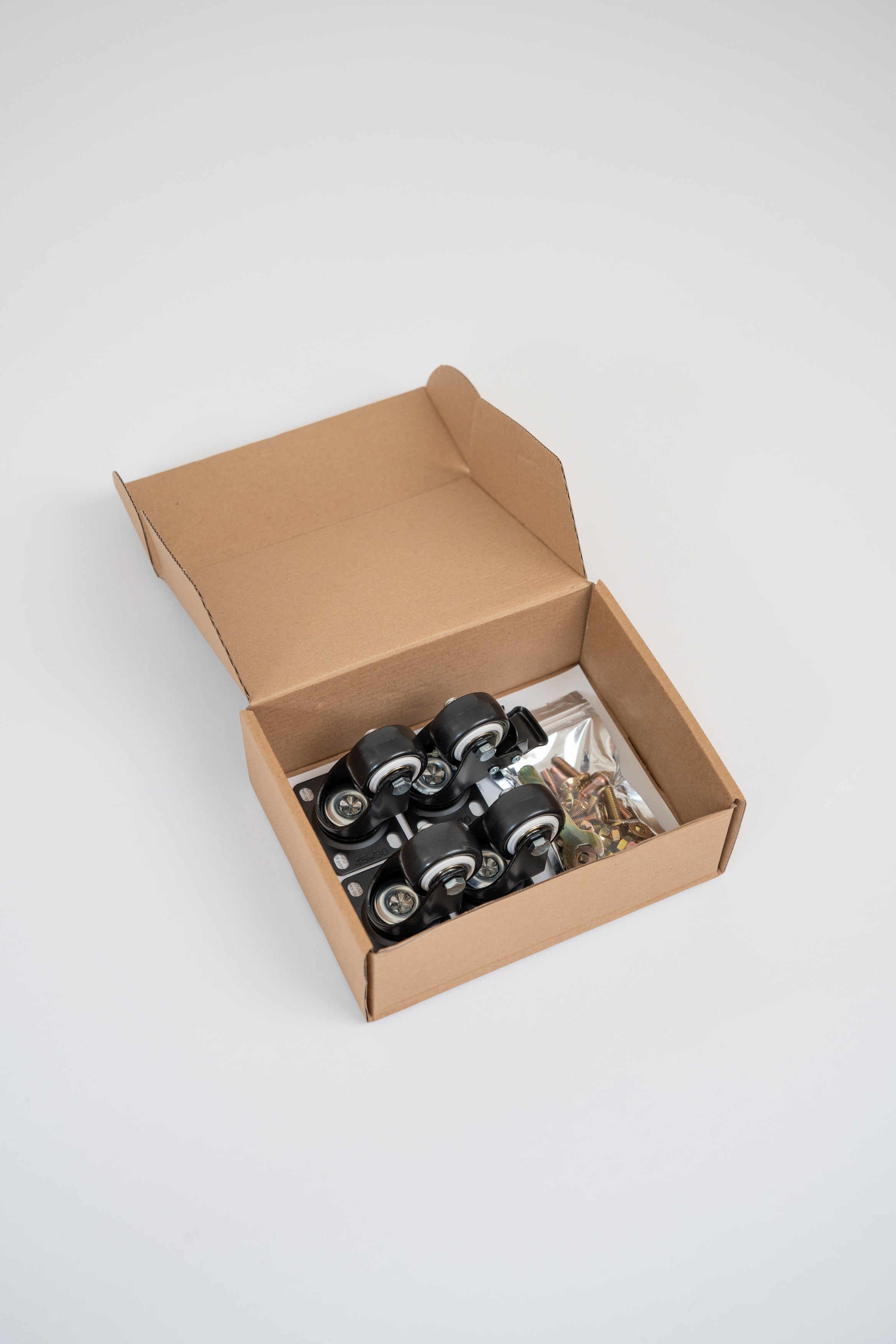 An open brown cardboard box containing four black caster wheels with metal axles and a small clear bag of hardware, photographed on a clean light gray studio background.