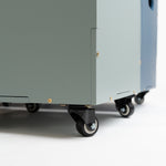 A close-up, low-angle shot of a large, industrial wheeled storage cabinet or utility cart in a muted sage green color, featuring black caster wheels and gold-colored mounting screws, set against a clean white studio background.