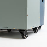 A close-up, low-angle shot of a large, industrial wheeled storage cabinet or utility cart in a muted sage green color, featuring black caster wheels and gold-colored mounting screws, set against a clean white studio background.