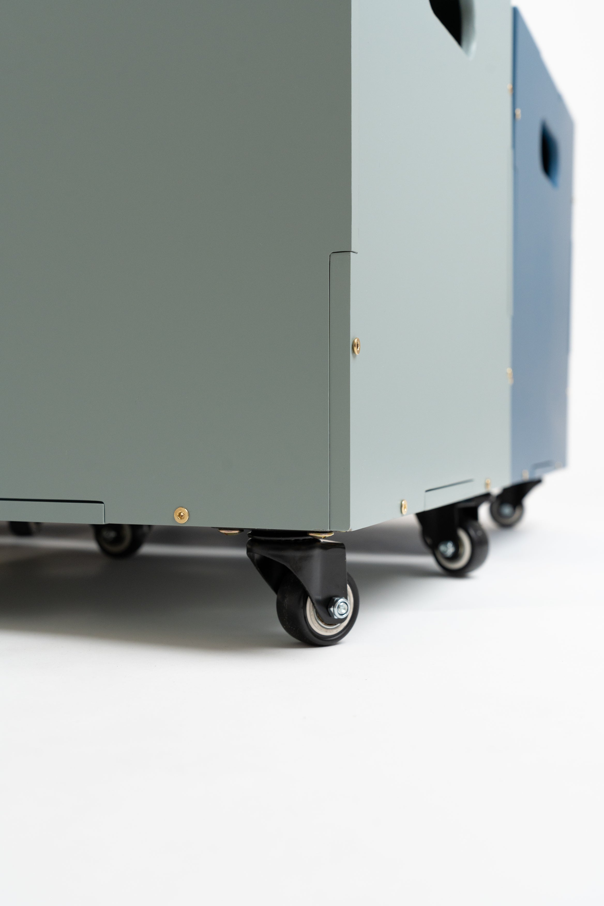 A close-up, low-angle shot of a large, industrial wheeled storage cabinet or utility cart in a muted sage green color, featuring black caster wheels and gold-colored mounting screws, set against a clean white studio background.