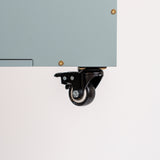 Close-up of a black caster wheel with a swivel bracket mounted under the edge of a light gray cabinet or panel, shown against a plain white background.
