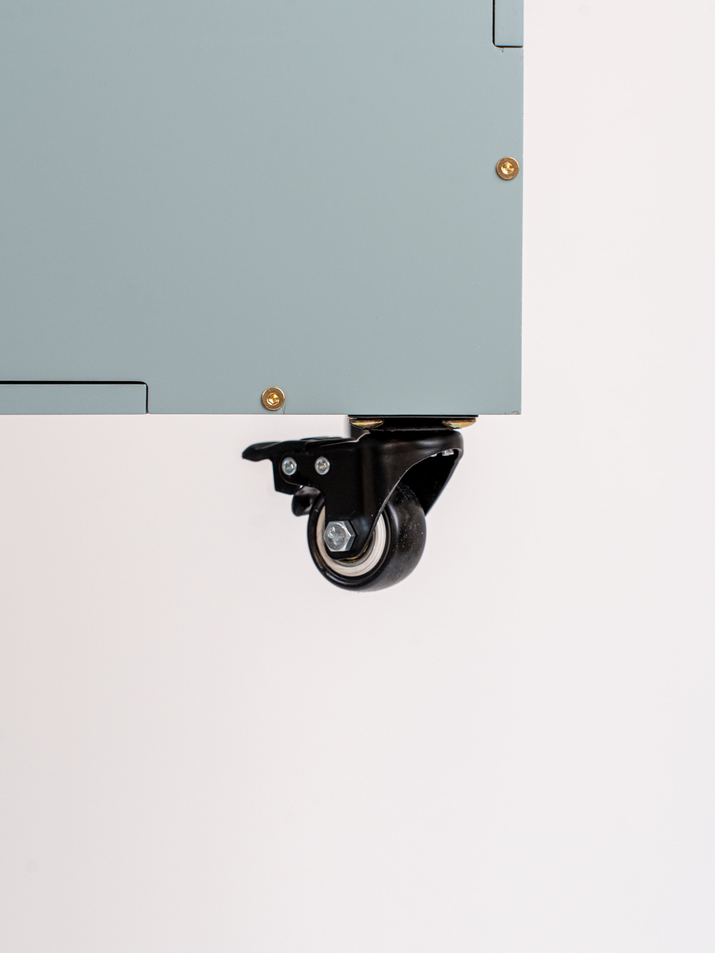Close-up of a black caster wheel with a swivel bracket mounted under the edge of a light gray cabinet or panel, shown against a plain white background.