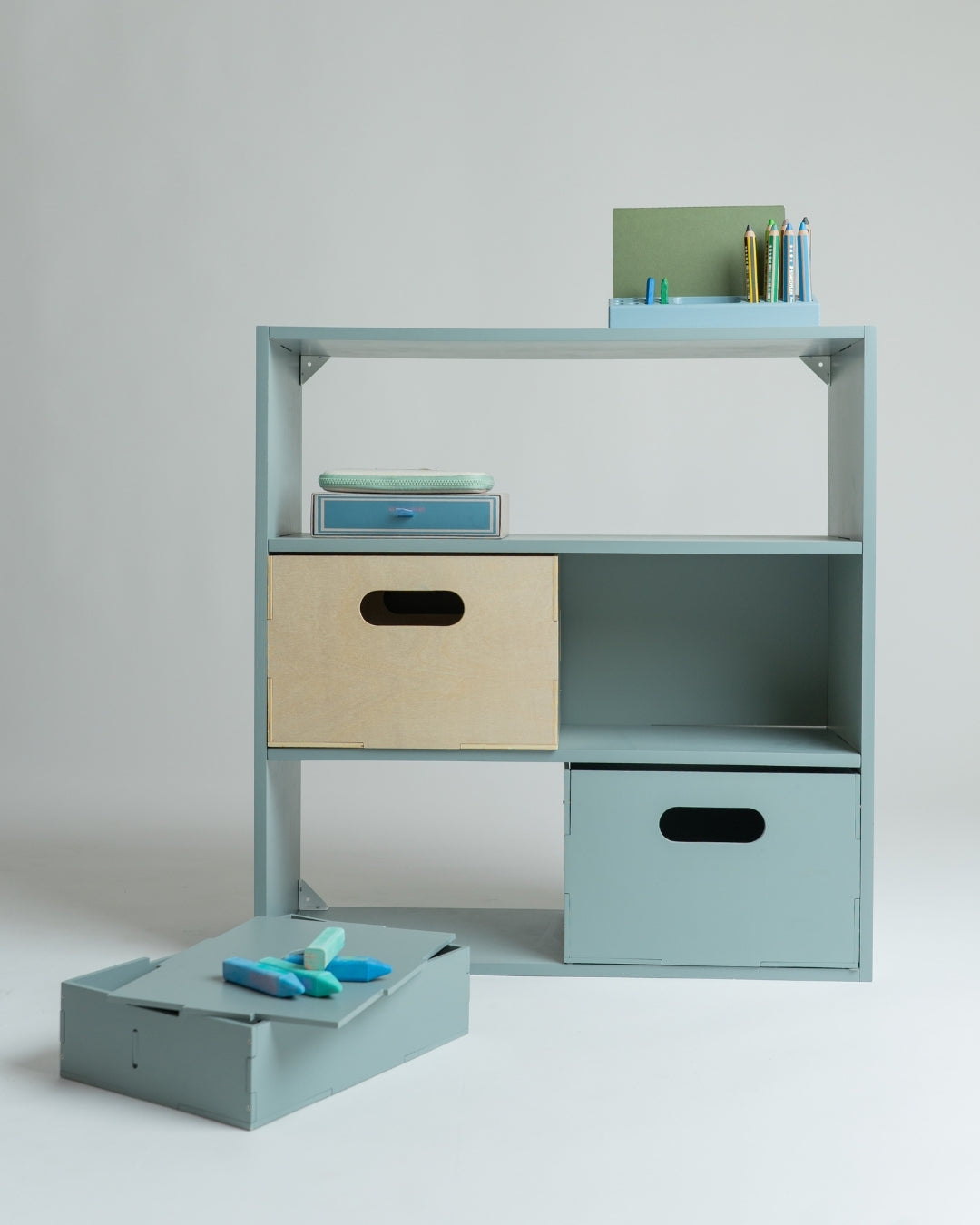 A light blue modular storage shelf unit with open shelves and two storage bins, shown on a neutral studio background with some books and toys as props.