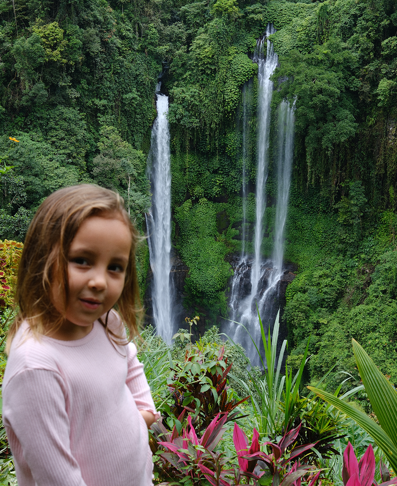 Child in a pink sweater standing in front of a waterfall surrounded by lush greenery