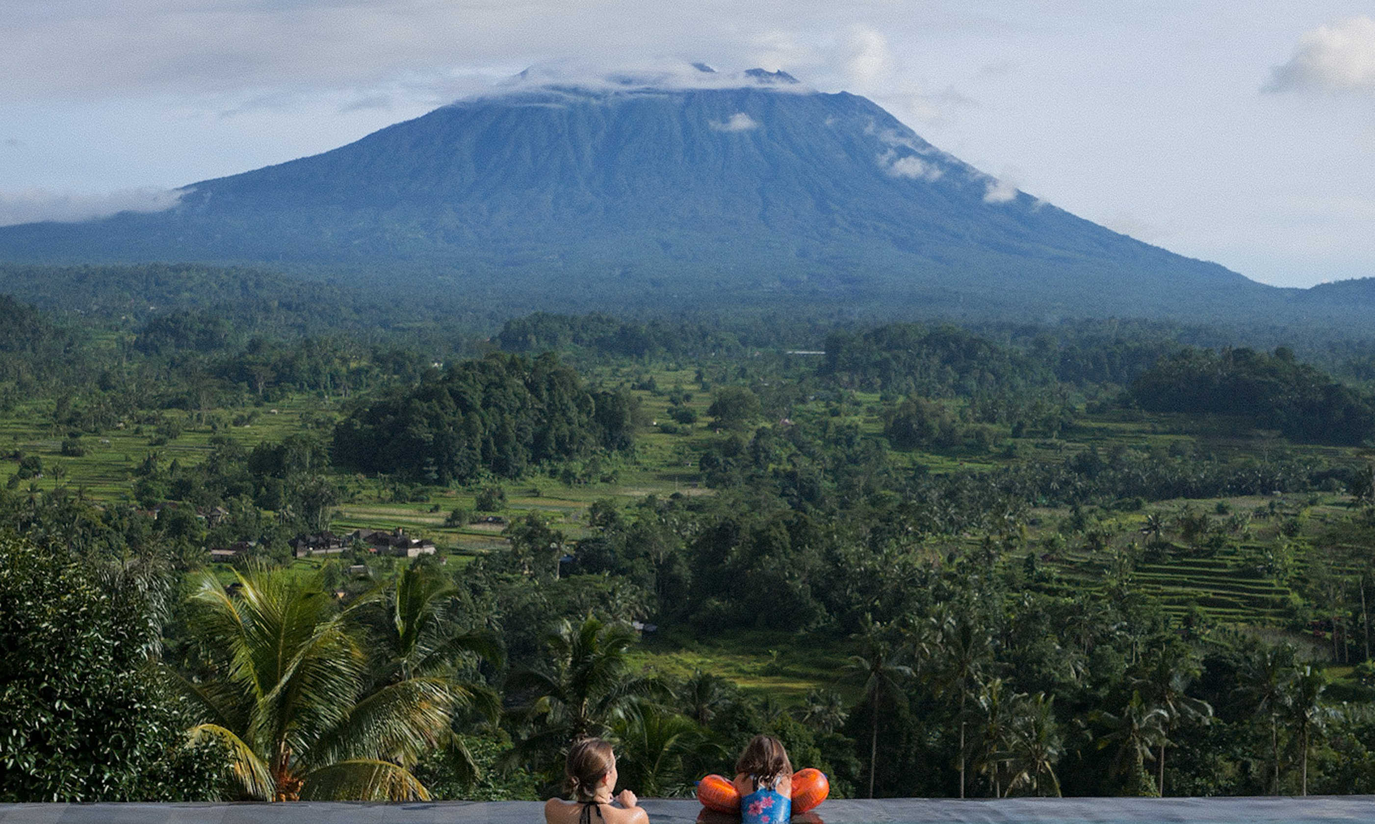 People in an infinity pool with a scenic view of a mountain and lush greenery.