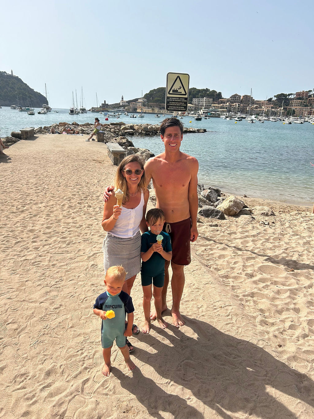 A family of four posing on a sandy beach with the sea and harbor in the background under a clear blue sky.
