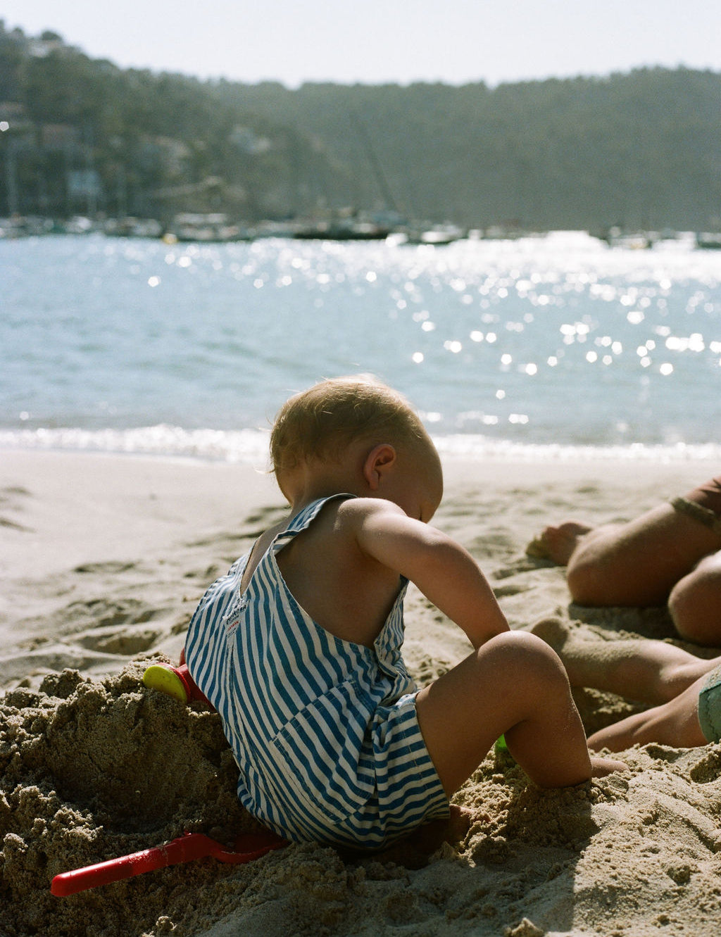 A toddler wearing a blue and white striped romper sits on a sandy beach next to a red plastic shovel, with the ocean and a tree-lined shore in the background.