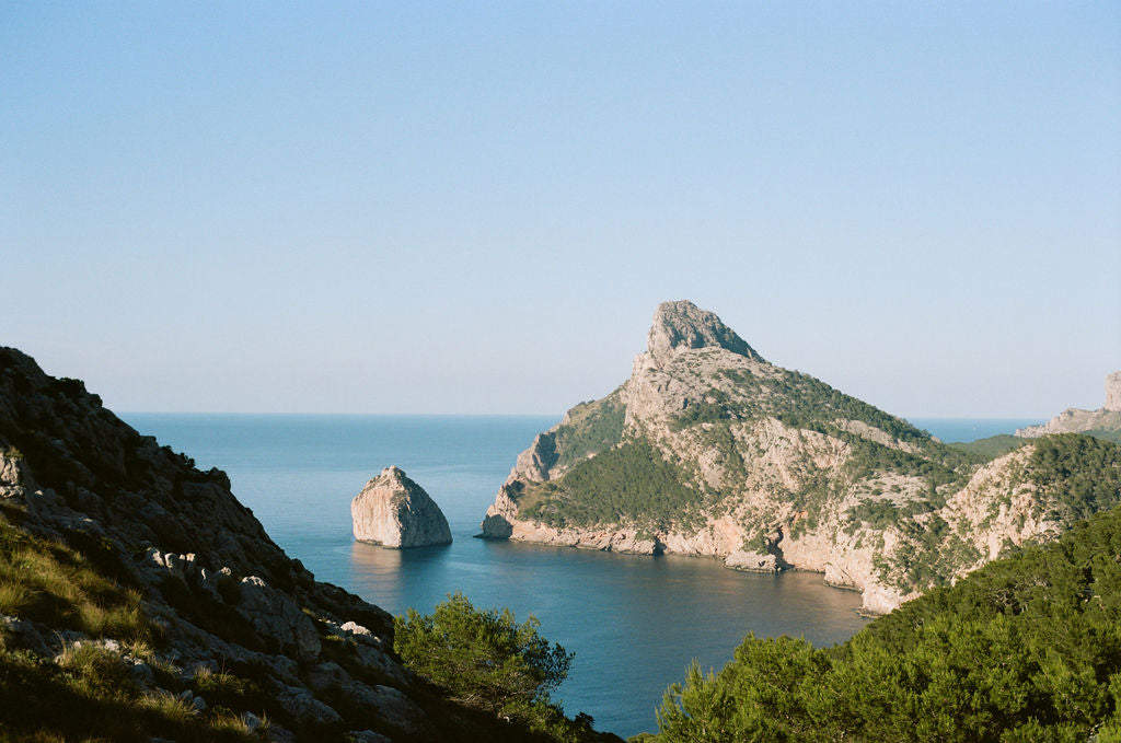 A scenic coastal landscape with rocky sea stacks rising from calm blue water under a clear pale blue sky, framed by green shrubs and sunlit hills in the foreground.
