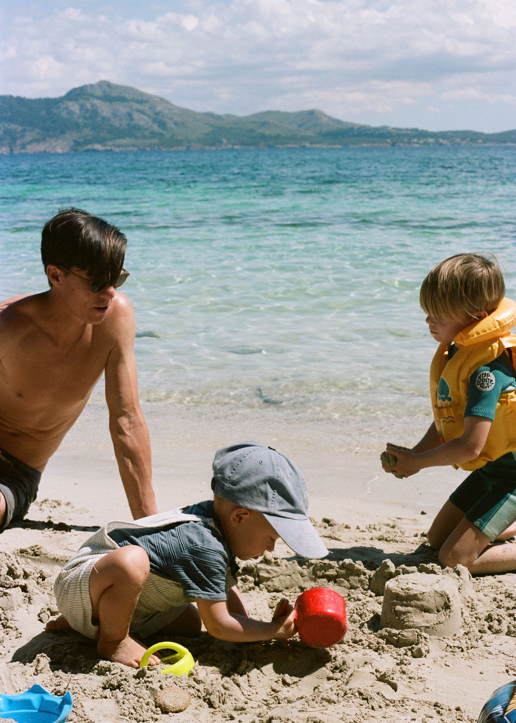 A family scene on a sandy beach with a man, a young boy building sandcastles, and a toddler in a yellow life vest. The turquoise sea and mountains are in the background.
