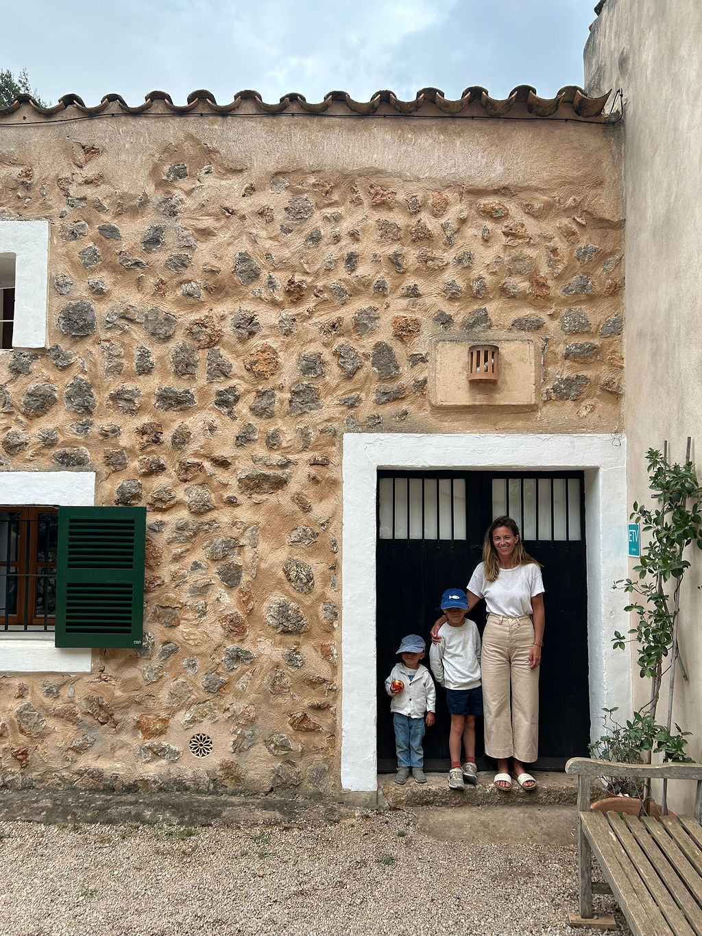 A woman and two children stand in the doorway of a rustic stone building with a terracotta tiled roof, white-trimmed window, and green shutters.
