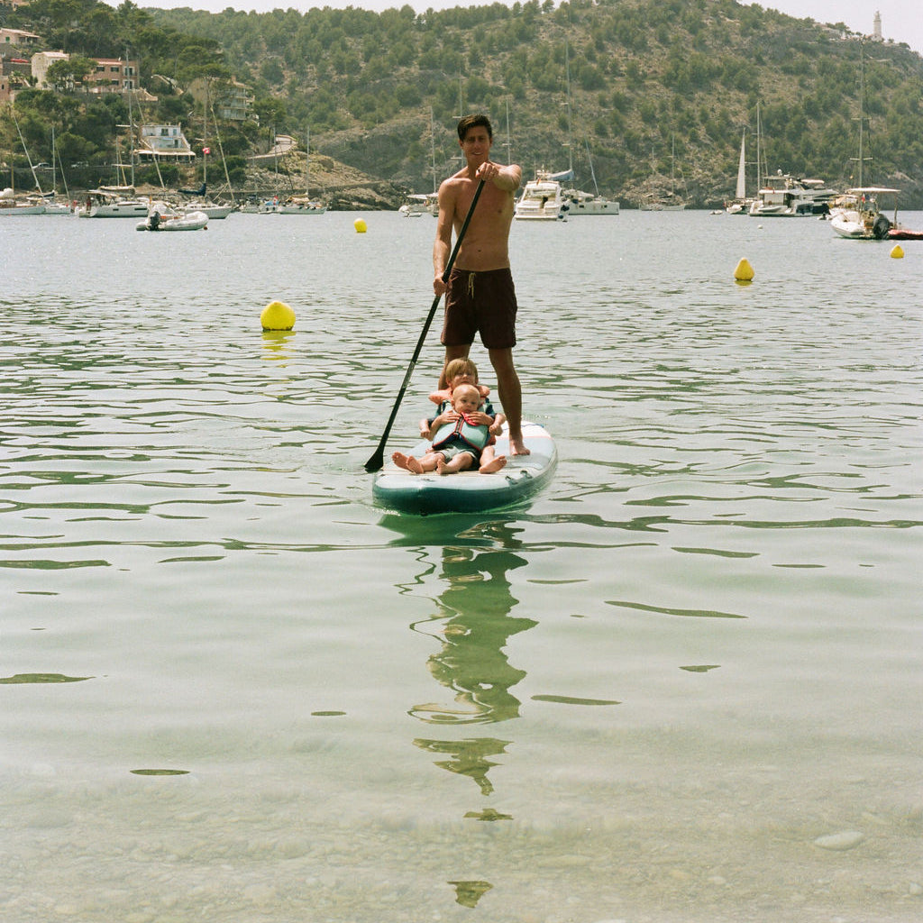 A man stands on a paddleboard in calm water while a child sits on his shoulders, with boats and a green hillside in the background.