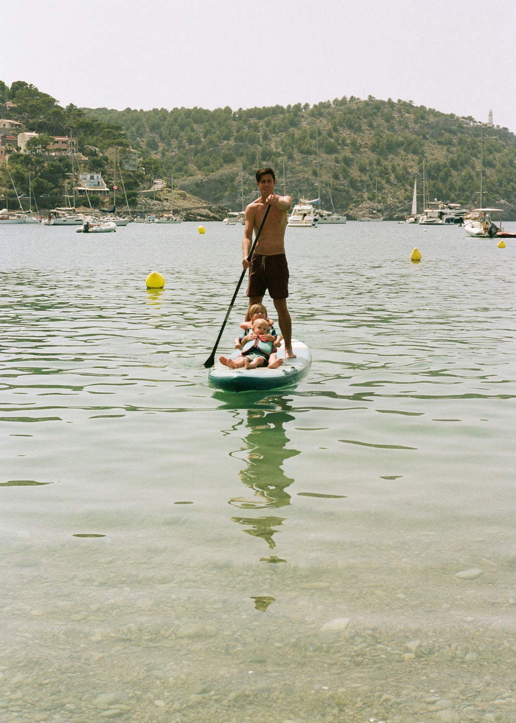 A man stands on a paddleboard in calm water while a child sits on his shoulders, with boats and a green hillside in the background.