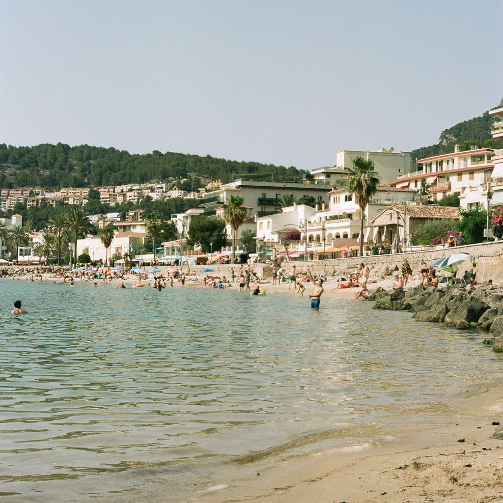 A scenic beach view with pale blue water, sandy shore, small beachgoers, buildings, palm trees, and a hillside background under a clear sky.