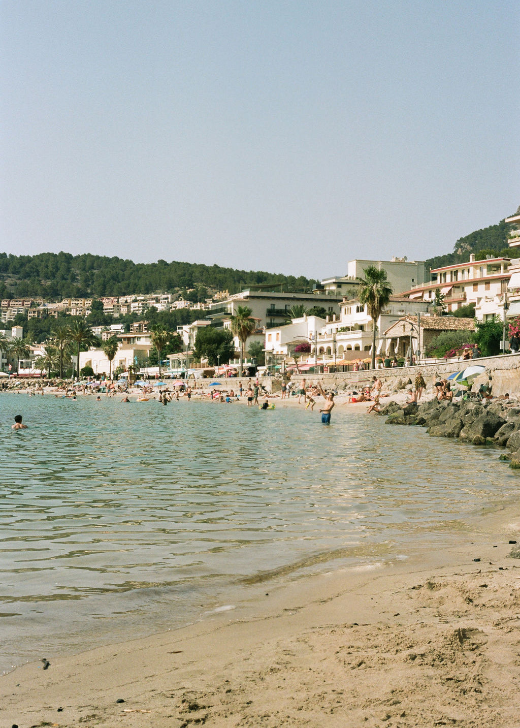 A scenic beach view with pale blue water, sandy shore, small beachgoers, buildings, palm trees, and a hillside background under a clear sky.