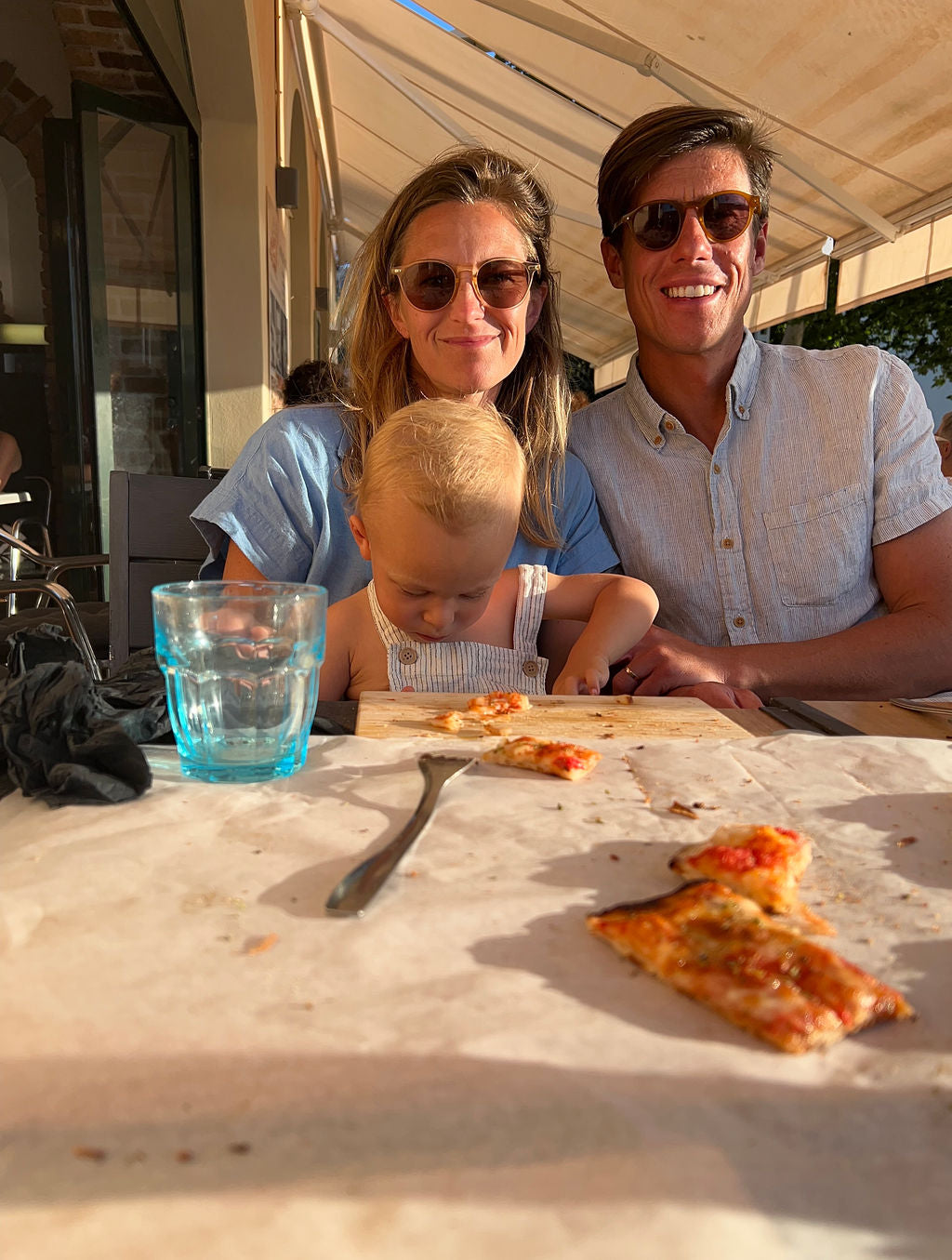 A family of three, including a man, a woman, and a young child, sitting at a table with pizza slices and a glass of water under a patio awning.