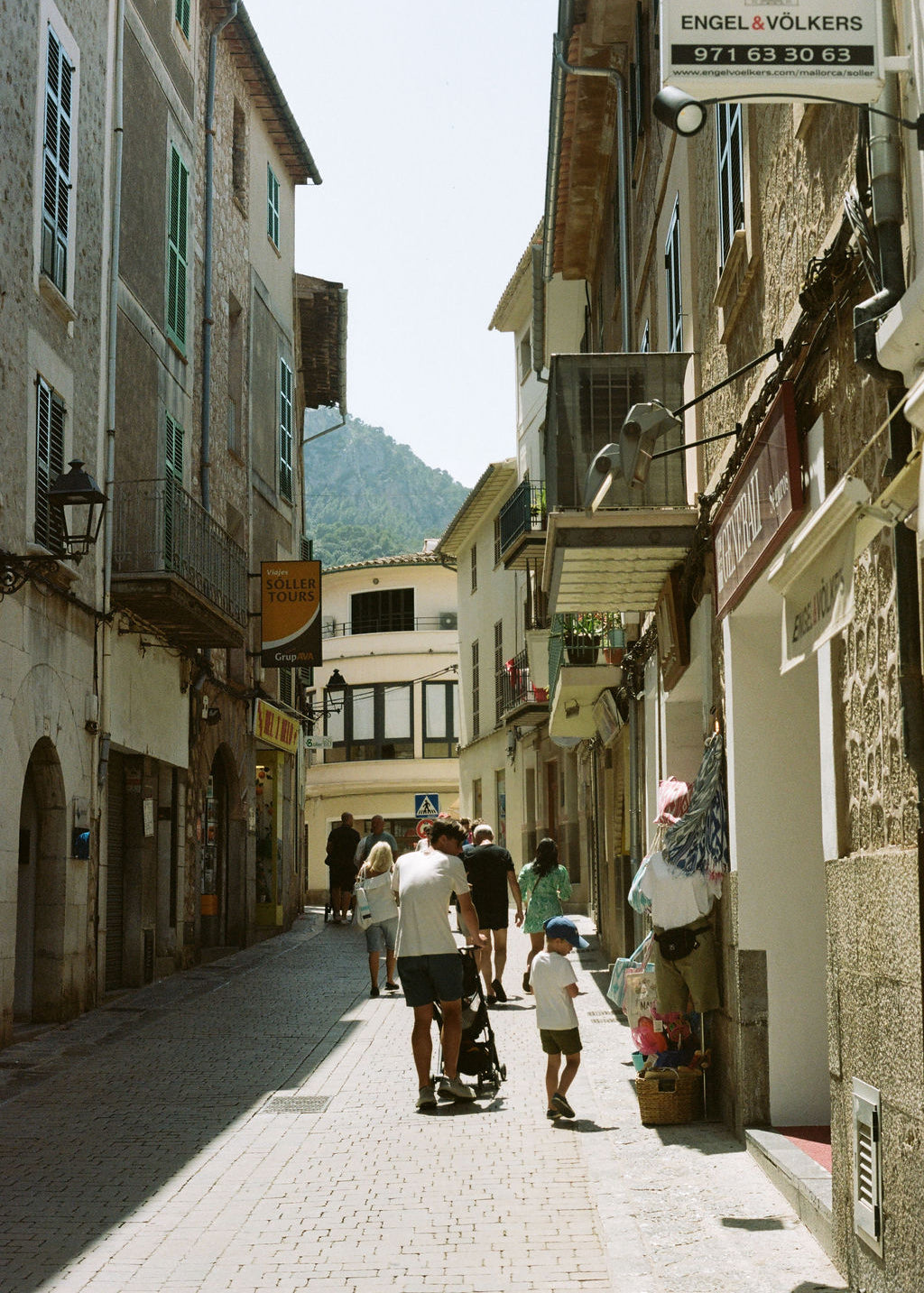 A sunlit cobblestone street in a historic European town with narrow buildings on both sides, people walking in the distance, and a mountain visible in the background. A hanging sign on the right reads Engel & Volkers.