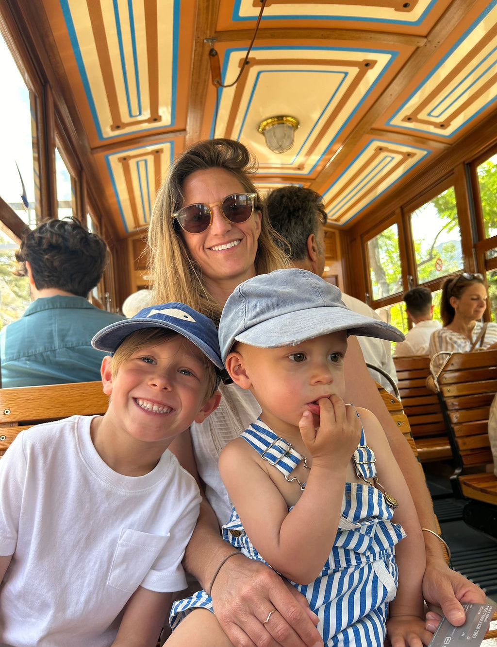 A family of three posing on a wooden trolley train, with a woman in sunglasses in the back, a young boy in a blue cap and white shirt in the front left, and a toddler in a blue and white striped top and cap in the front right.