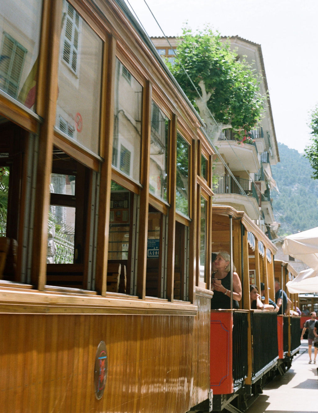 A vintage wooden cable car with large windows travels along a street beside buildings and greenery.