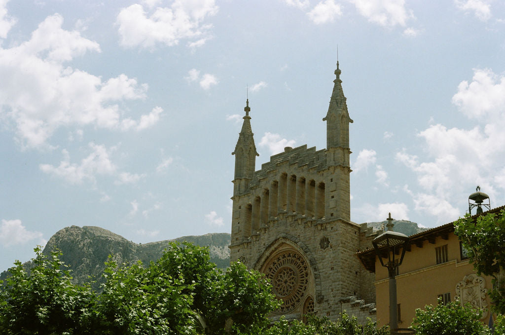 A scenic photograph of a historic stone cathedral with two spires, set against a blue sky with white clouds, green trees in the foreground, and distant mountains.